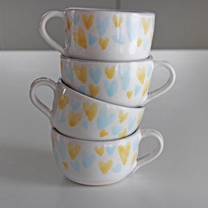 May include: A stack of four white ceramic mugs with blue and yellow heart patterns.