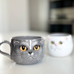 May include: Two ceramic mugs shaped like cat faces. The mug on the left is grey with orange eyes and a white stripe on its nose. The mug on the right is white with yellow eyes.