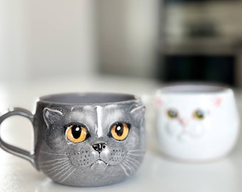May include: Two ceramic mugs shaped like cat faces. The mug on the left is grey with orange eyes and a white stripe on its nose. The mug on the right is white with yellow eyes.