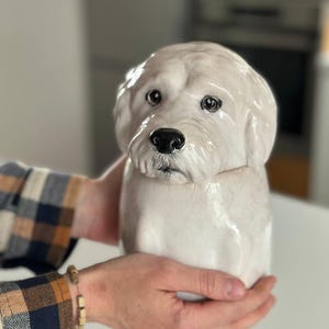 May include: A ceramic dog figurine featuring a cream-coloured coat with a glossy finish. The sculpture depicts a dog head with detailed facial features, including dark eyes and a black nose, transitioning into a cylindrical body base.