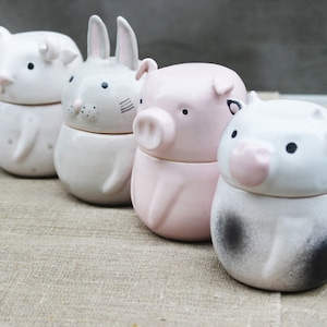 Może przedstawiać: Cztery ceramiczne pojemniki w kształcie zwierząt. Pojemniki są białe, różowe i szare, z czarnymi oczami i różowymi akcentami. Zwierzęta obejmują niedźwiedzia, królika i dwa prosięta. Pojemniki są ułożone w rzędzie na neutralnej powierzchni.