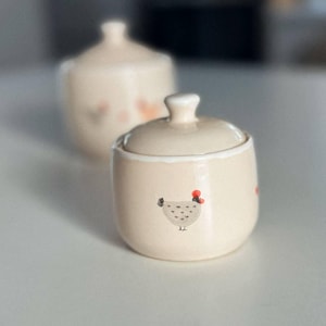 Op de afbeelding: Twee crèmekleurige keramische suikerpotten met deksels. De voorste pot heeft een handgeschilderd kippenontwerp met een rode kam. De potten staan op een wit oppervlak, waarbij de tweede pot enigszins onscherp is op de achtergrond.