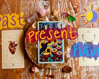 May include: Tarot card reading with three cards laid out on a wooden surface. The cards are labeled "Past", "Present", and "Future". The card for the past shows a heart with a needle and thread. The card for the present shows a black bird on a branch with sunflowers and red flowers. The card for the future shows a lamp with three flames.