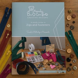 May include: A collection of sewing supplies, including zippers, buttons, and a guide to zips and fasteners. The guide is titled "Family Comforts Creative Designs" and features a bear logo. Various colors of zippers and buttons are scattered around the guide.