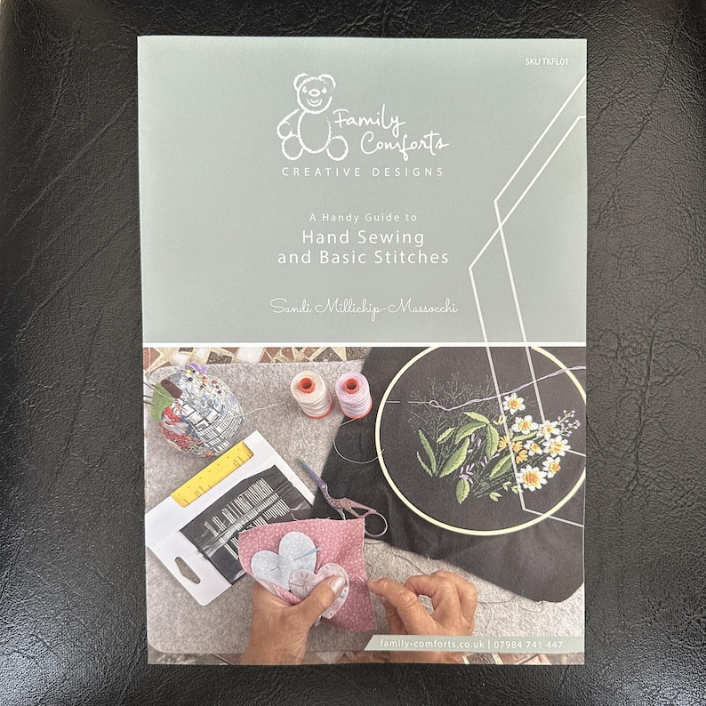 May include: A book titled "Family Comforts Creative Designs" with the subtitle "A Handy Guide to Hand Sewing and Basic Stitches" is displayed. The image shows sewing supplies, including thread spools, needles, and an embroidery hoop with floral designs.