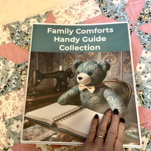 May include: A binder titled "Family Comforts Handy Guide Collection" featuring a teddy bear with a bow tie, a vintage sewing machine, and an open notebook. The binder is held by a hand with a gold ring and dark red nail polish.