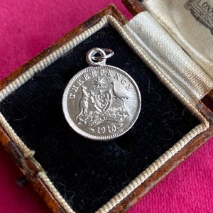 May include: A silver pendant featuring a coin with the text "THREE PENCE" and the year "1910". The pendant is displayed in a vintage, brown leather jewelry box with a black velvet interior and a cream-colored satin lining.
