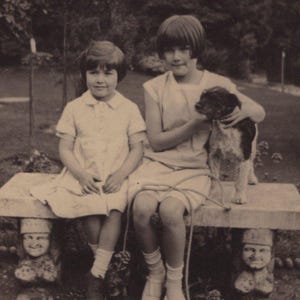 Op de afbeelding: Een vintage zwart-witfoto van twee kinderen die op een stenen bank met gebeeldhouwde steunen zitten. Eén kind houdt een kleine hond aan de lijn. De kinderen dragen lichtgekleurde jurken en sokken, en de scène speelt zich buiten af.