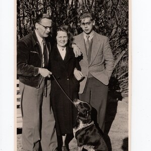 May include: A black and white photo of a family with a dog. A man in a dark jacket and a woman in a coat stand with a young man in a suit jacket. The dog is a black and white spotted breed.