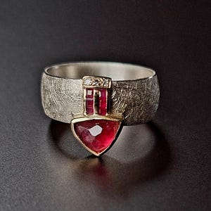 Könnte beinhalten: Ein silberner Ring mit einer strukturierten Band und einem roten Edelstein, der in eine goldene Fassung gefasst ist. Der Edelstein hat die Form eines Herzens mit einem kleineren Diamanten darüber.