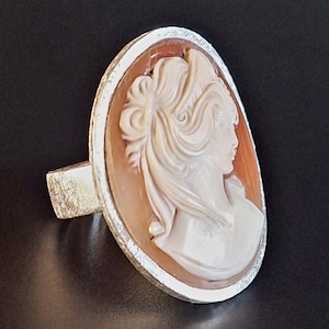 Könnte beinhalten: Ovale Cameo-Ring mit dem Profil einer Frau mit fließendem Haar, in cremefarbenem Relief vor hellorangefarbenem Hintergrund geschnitzt. Der Cameo ist in einem silberfarbenen Metallring gefasst.