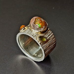 Könnte beinhalten: Ein Silberring mit strukturiertem Band und drei Edelsteinen. Der größte Edelstein ist oval mit orangefarbenen, braunen und grünen Farbtönen. Zwei kleinere Edelsteine sind an den Seiten des Rings gesetzt.