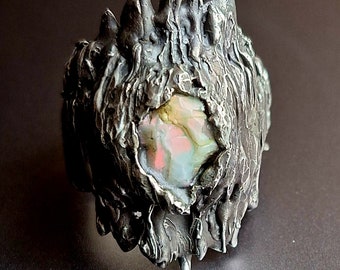 May include: A silver ring with a textured, organic design. The ring features a large, iridescent gemstone set in the center.