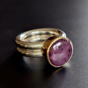 Puede incluir: Un anillo con una banda de plata pulida y una piedra preciosa morada de corte cabujón, engastada en un bisel de tono dorado. La piedra muestra inclusiones naturales y una superficie lisa y abovedada que contrasta con el brillo de la plata.