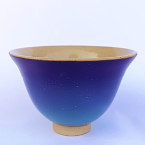 Può includere: Una ciotola in ceramica con bordo svasato, caratterizzata da una finitura sfumata liscia che passa da un blu reale intenso nella parte superiore a un azzurro cielo più chiaro alla base. L'interno e il piccolo piede circolare sono beige.