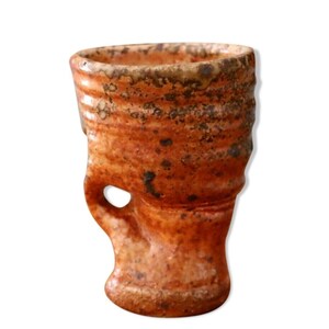 Può includere: Una tazza in ceramica fatta a mano con una forma organica unica. La tazza è principalmente arancione con macchie marroni scure e un piccolo manico con un foro. La superficie strutturata e i toni della terra suggeriscono un'estetica rustica e artigianale.