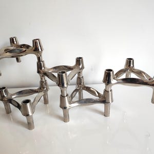 Peut inclure: Ensemble de bougeoirs en métal argenté. Chaque bougeoir présente un design sculptural unique avec trois ouvertures pour bougies et une base tripode. Les bougeoirs sont disposés sur une surface blanche.