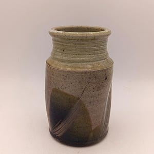 Puede incluir: Un jarrón cilíndrico de cerámica con un acabado moteado en beige y gris. El cuello superior presenta estrías horizontales, mientras que el cuerpo inferior muestra patrones de pinceladas en verde oscuro y marrón con sutiles hendiduras verticales.