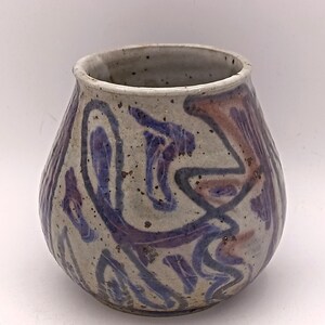 Può includere: Un vaso in ceramica fatto a mano, dalla forma bombata e con un'ampia apertura. Il vaso è principalmente bianco sporco con disegni astratti blu e rosso-marrone. La superficie ha un aspetto strutturato, che aggiunge fascino rustico.