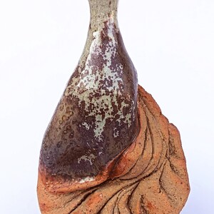 Peut inclure: Un vase en céramique avec un col étroit et une base large et texturée. La partie supérieure présente un vernis brillant tacheté de brun et de beige, tandis que la section inférieure est en argile rouge-brun non émaillée avec des rainures.