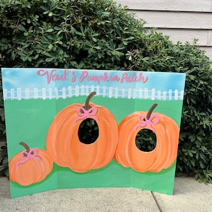 May include: A painted cardboard cutout of three pumpkins with pink bows. The largest pumpkin has a hole cut out of it. The cutout is labeled "Vail's Pumpkin Patch".