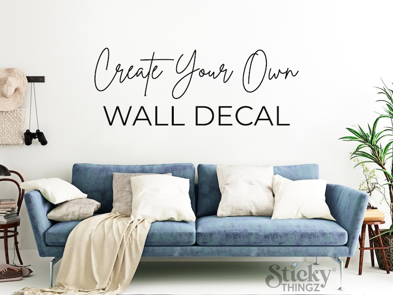 May include: A blue sofa with white pillows and a beige throw blanket. The wall above the sofa has the text "Create Your Own WALL DECAL" in black script. A decorative wall hanging and a plant are visible.