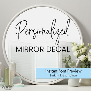 May include: A round white mirror with the words "Personalized MIRROR DECAL" in black script. A light blue rectangle at the bottom of the mirror reads "Instant Font Preview Link in Description". A vase of white flowers is to the right of the mirror.