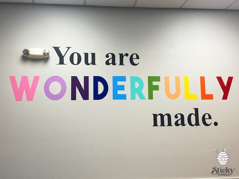 May include: A wall decal with the words "You are WONDERFULLY made" in black and rainbow colors. The word "WONDERFULLY" is in pink, purple, navy, blue, green, orange, yellow, and red. A white emergency light is mounted on the wall.