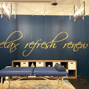 May include: A massage table with a blue surface is centered in front of a dark blue wall. The wall features gold script that reads "relax refresh renew." A wooden cabinet with white cubbies sits behind the table.