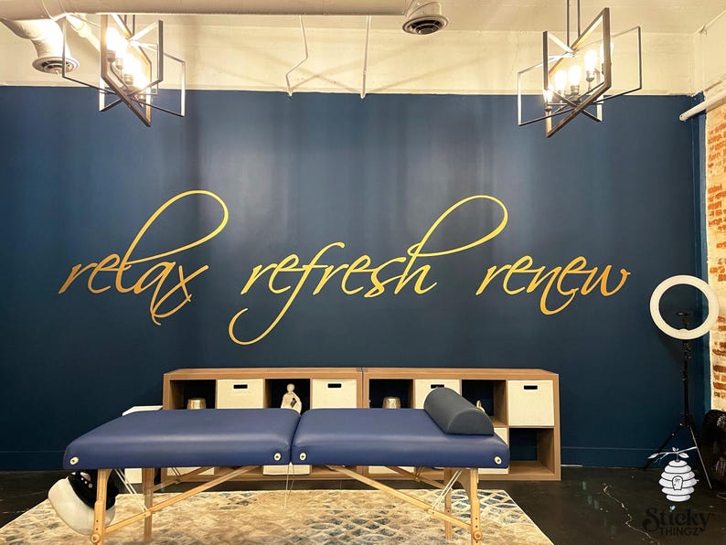 May include: A massage table with a blue surface is centered in front of a dark blue wall. The wall features gold script that reads "relax refresh renew." A wooden cabinet with white cubbies sits behind the table.