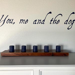 May include: A wooden shelf with six navy blue candles. Above the shelf, the phrase "You, me and the dog" is written in a dark blue, cursive font on a white wall. The shelf is placed on a gray cabinet.