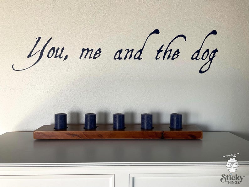May include: A wooden shelf with six navy blue candles. Above the shelf, the phrase "You, me and the dog" is written in a dark blue, cursive font on a white wall. The shelf is placed on a gray cabinet.