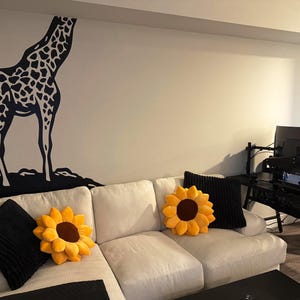 May include: A living room scene featuring a white sectional sofa adorned with sunflower-shaped pillows and black throw pillows. A large black and white giraffe wall decal is on the wall, adding a whimsical touch to the decor.