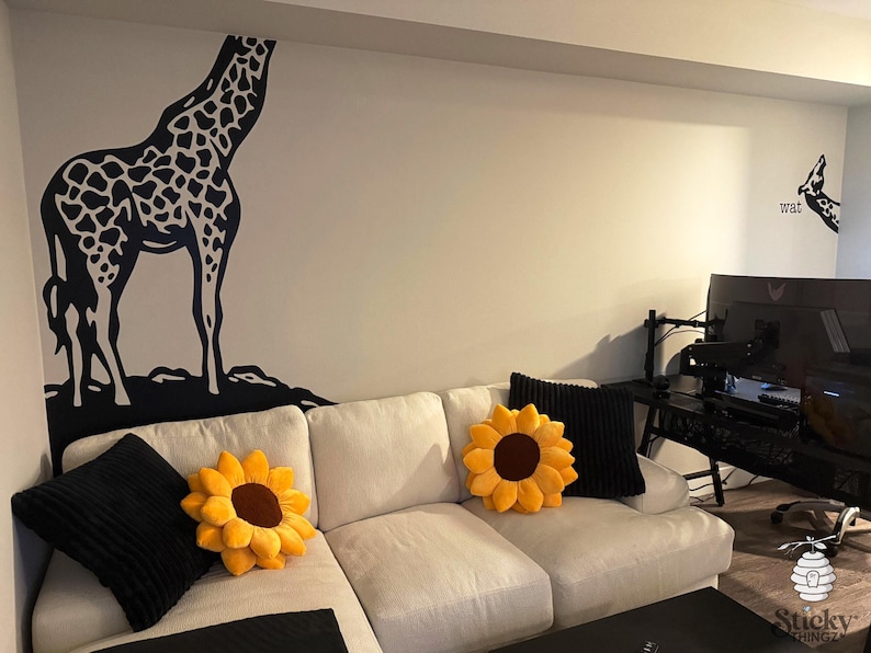 May include: A living room scene featuring a white sectional sofa adorned with sunflower-shaped pillows and black throw pillows. A large black and white giraffe wall decal is on the wall, adding a whimsical touch to the decor.