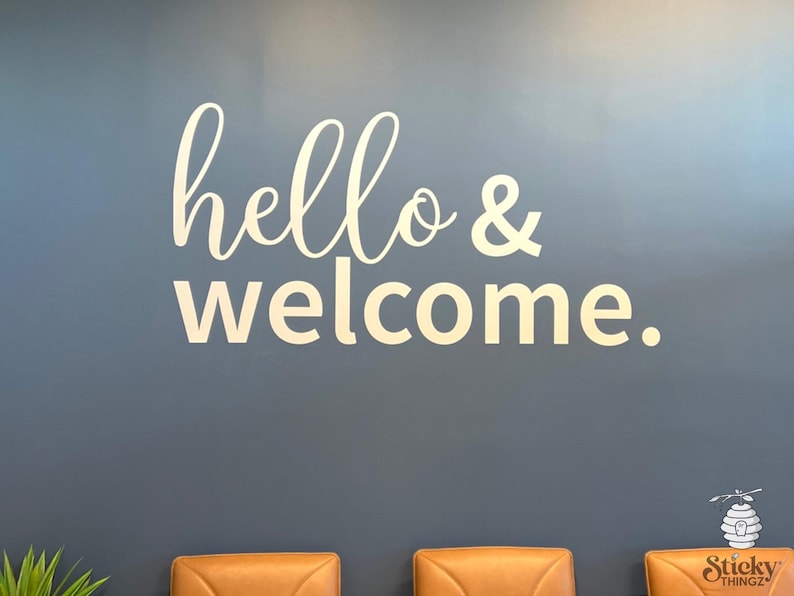 May include: A wall decal with the words "hello & welcome" in a cursive font. The text is in a light color and is placed against a dark blue wall. The decal is a decorative element for a home or business.