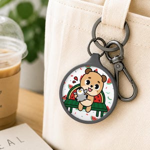 May include: A circular keychain featuring a cartoon bear holding a small penguin while sitting on a green bench. The background pattern includes watermelon slices. The keychain has a dark gray metal frame and a matching lobster clasp attachment.