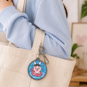 May include: A circular keychain featuring a cartoon pink rabbit wearing black sunglasses while floating in a blue swimming pool on a pink flamingo inner tube. The keychain is attached to a silver metal clasp and hangs from a beige canvas tote bag.