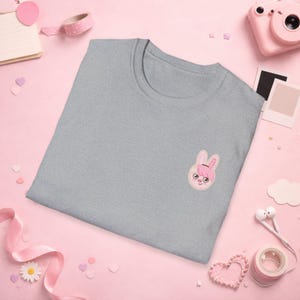 Puede incluir: Camiseta gris claro con un bordado de una cara de conejo con orejas rosas, un lazo rosa y ojos redondos. La camiseta está hecha de material suave y es adecuada para uso casual.