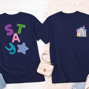 Könnte beinhalten: Zwei marineblaue T-Shirts. Eines zeigt das Wort "STAY" in bunten Buchstaben und Formen im Patchwork-Stil. Das andere hat eine Hausgrafik mit dem Wort "STAY" auf der Vorderseite. Die Hemden werden auf einer hellen Oberfläche präsentiert.