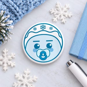 May include: A circular button featuring a cartoon face with a light blue complexion, wearing a white beanie with blue snowflake patterns. The character has large dark blue eyes, rosy cheeks, and a blue pacifier in its mouth, rendered in a flat style.