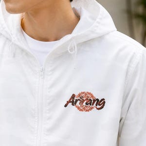 Peut inclure: Veste à capuche blanche avec un motif camouflage. La veste est dotée d'une fermeture éclair intégrale et d'une capuche à cordon de serrage. Le mot "Arirang" est brodé sur la poitrine en marron et orange.