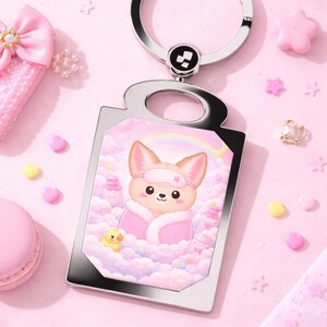 May include: A silver-tone keychain with a rectangular charm featuring a cartoon fox in a pink robe and eye mask, relaxing in a bubble bath. The background is a soft pink with a rainbow. The keychain is surrounded by pink accessories.
