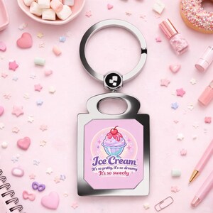 May include: A silver keychain with a rectangular charm featuring an ice cream graphic and the text "Ice Cream". The background is a pastel pink with scattered heart and star-shaped decorations, and other pink accessories.