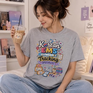 May include: A heather gray short-sleeve t-shirt featuring a colorful graphic print that reads Korean EMS Tracking Specialist. The design includes illustrations of shipping boxes, a laptop, a smartphone, a coffee cup, and a checklist.