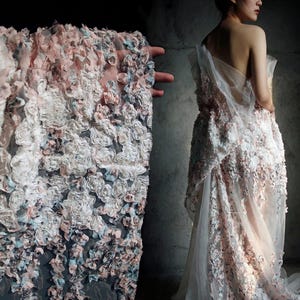 Könnte beinhalten: Ein weißes Brautkleid mit floralen Verzierungen in Rosa- und Blautönen. Das Kleid hat einen fließenden Rock und einen transparenten Überzug, was einen romantischen und ätherischen Look erzeugt. Der Stoff ist mit detaillierten floralen Applikationen strukturiert.