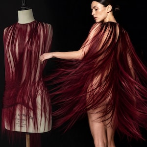 Puede incluir: Una prenda de color burdeos profundo construida con largos mechones fluidos similares al cabello. La pieza está drapeada sobre un maniquí con el texto 'STOCKMAN PARIS 50497' visible en el torso, mientras que también la usa una persona como capa.