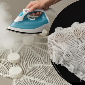 May include: A blue and white steam iron being used on a white textured fabric. The iron is held by a person's hand. The fabric has a pattern of raised, circular elements.