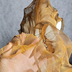 May include: Close-up of a textured, layered fabric in shades of gold and brown. The fabric appears to be draped and folded, creating a sense of depth and dimension. The material has a ribbed pattern.