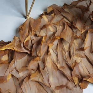May include: A flowing, decorative fabric in shades of brown and gold, with a layered, textured appearance. The fabric appears to be draped, with a light, airy quality. The material is likely silk or a similar delicate textile.