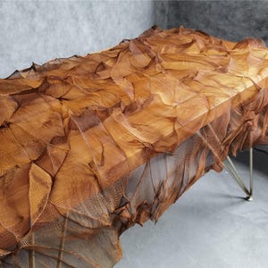 May include: A unique, sculptural table with a textured, layered surface in shades of brown and copper. The table has a rectangular top and is supported by thin, silver-colored legs. The design evokes a sense of organic forms and artistic craftsmanship.